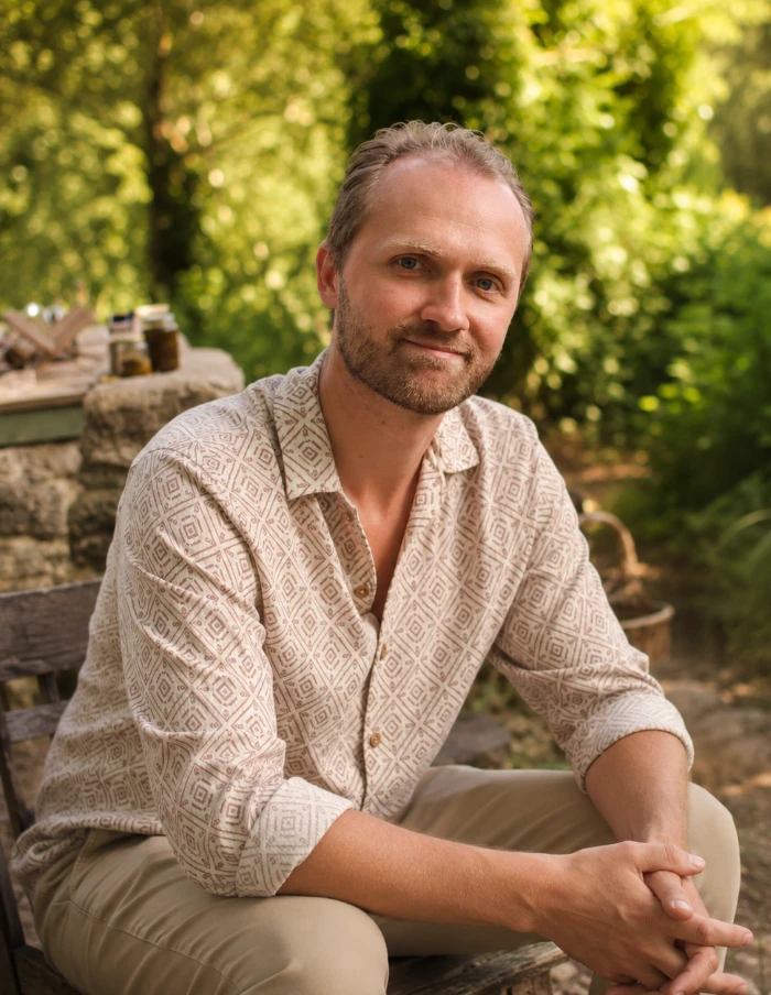 Serapis Light relaxed profile photo A man with short light brown hair and a trimmed beard sits outdoors on a chair, wearing a beige patterned shirt and light pants, with green foliage and sunlight in the background.